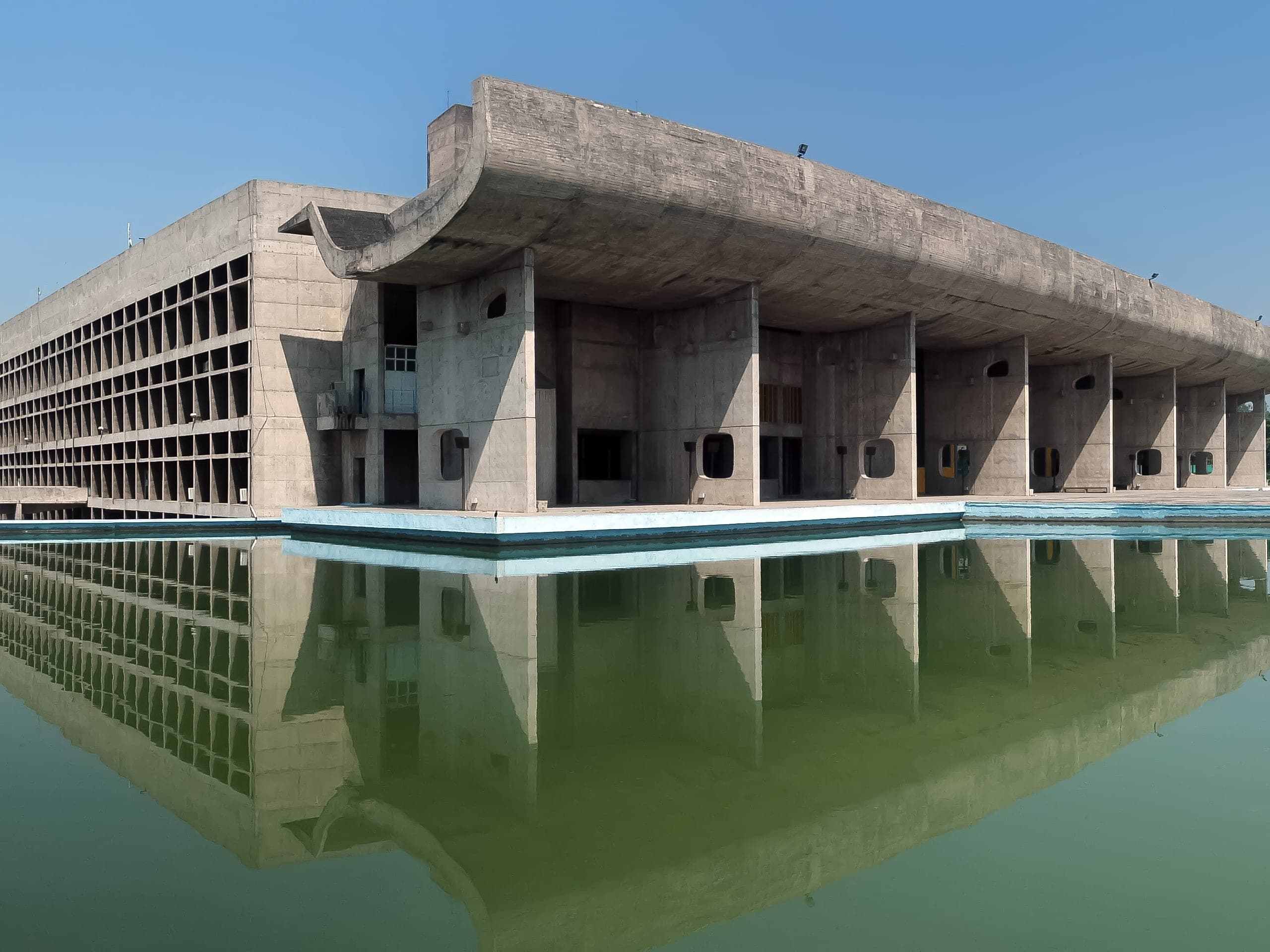 A concrete parliament building with a sweeping curved roof, reflected in a long pool