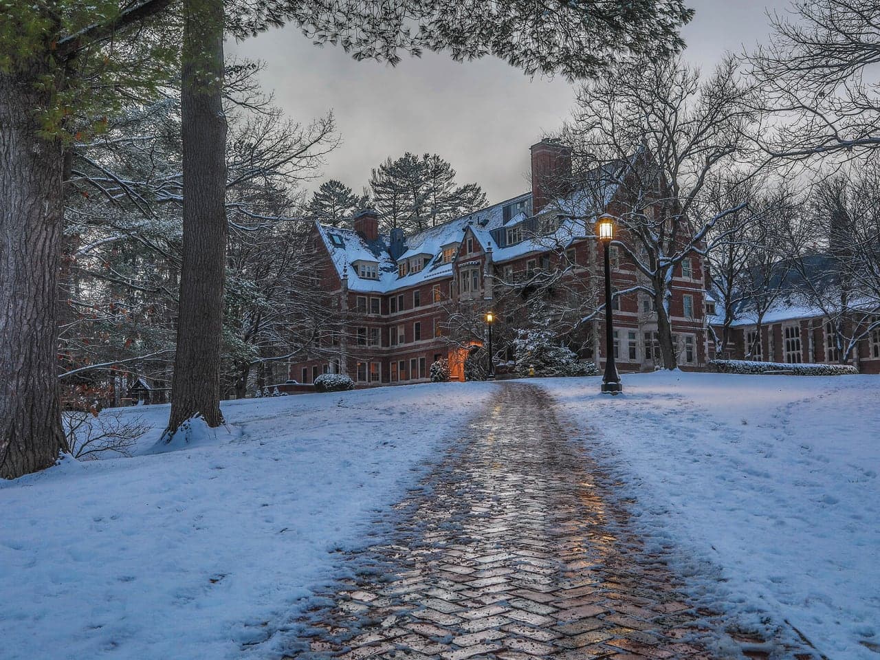 A snowy brick path at St. Paul’s School leading to a brick building, with lamps lit at dusk