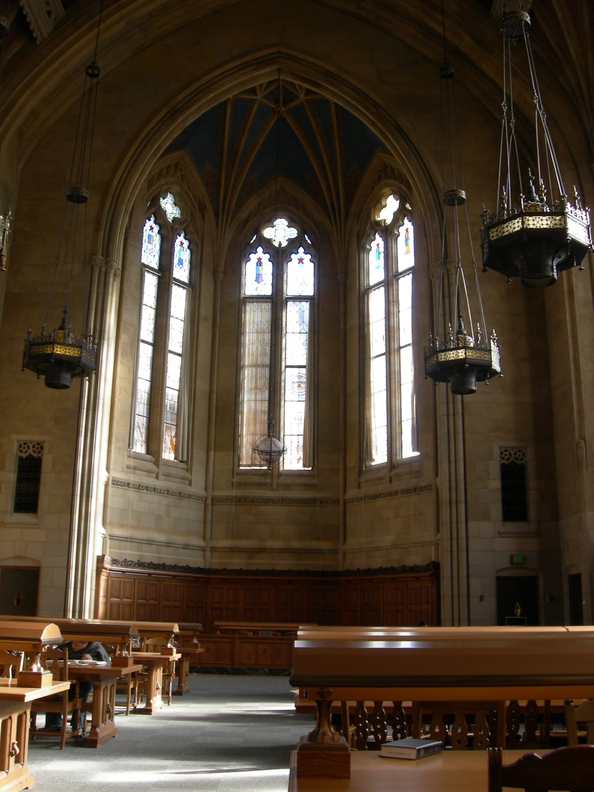 A tall Gothic reading room with stained-glass windows, hanging lamps, and long wooden study tables