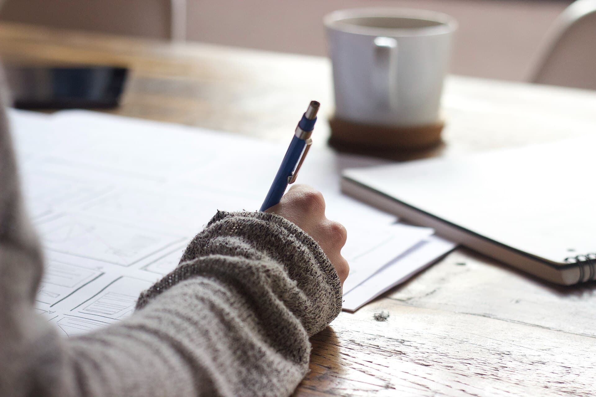A hand writing notes with a blue pen at a wooden table, beside a mug and a notebook